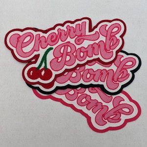 Op de afbeelding: Drie roze en witte strijklabels met de tekst "Cherry Bomb" in een retro-lettertype. De labels zijn op elkaar gelegd.