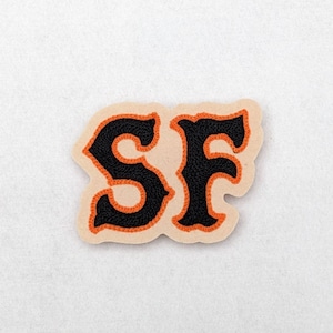 May include: A beige felt patch with an orange outline and black felt letters spelling "SF".