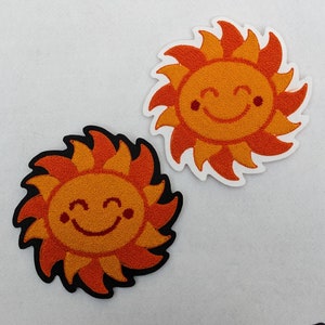 May include: Two felt appliques of smiling suns with orange and red rays. One sun is black and the other is white.