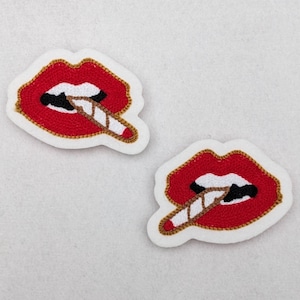May include: Two red embroidered lips with a white and brown cigarette sticking out of them. The lips are outlined in gold thread and are on a white background.