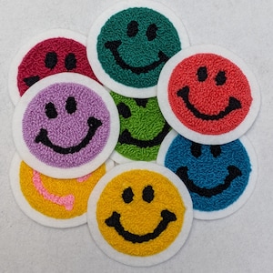 Peut inclure: Neuf patchs brodés en forme de smiley de différentes couleurs, notamment vert, rouge, violet, jaune et bleu. Chaque patch est circulaire et présente un contour blanc avec un smiley noir.