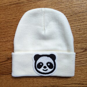 Può includere: Berretto color crema con bordo ripiegato. Una toppa con la faccia di un panda in bianco e nero è centrata sul davanti. Il cappello è realizzato in morbido materiale a maglia.
