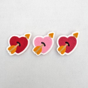 May include: Three felt heart-shaped patches with arrows through them. The hearts are red, pink, and burgundy. The arrows are gold.