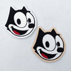 Könnte beinhalten: Zwei gestickte Aufnäher mit einem Cartoon-Katzenkopf, der ein breites Grinsen und große Augen hat. Die Katze ist schwarz-weiß mit einem roten Mund. Ein Aufnäher ist auf weißem Grund, der andere auf braunem Grund.