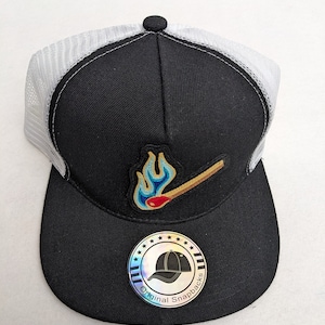 May include: Black and white trucker hat with a mesh back. The hat has a patch of a lit match with a blue and yellow flame. The hat also has a black and white sticker with the text "Original Snapbacks".