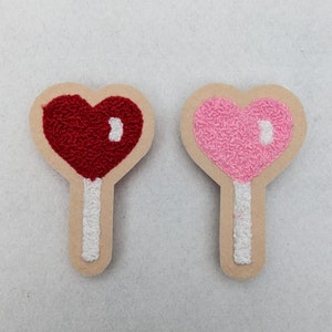 May include: Two embroidered felt heart-shaped lollipops, one red and one pink, with white stick details.