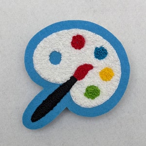 May include: A blue felt patch with a white paint palette and a black and red paintbrush. The palette has five paint wells with red, blue, yellow, green, and white paint.