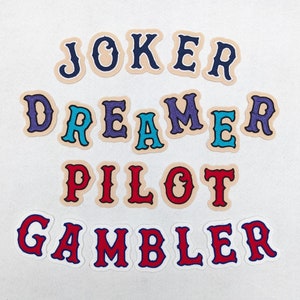 Pode incluir: Um conjunto de nove patches termocolantes coloridos com as palavras "JOKER", "DREAMER", "PILOT" e "GAMBLER" em uma variedade de cores e fontes.