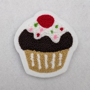 May include: A brown and white embroidered cupcake with a red cherry on top. The cupcake is decorated with white frosting and colorful sprinkles.