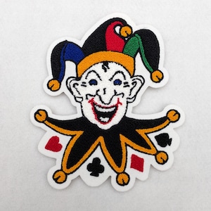 May include: An embroidered patch depicting a smiling jester in a black and yellow outfit and a multi-coloured hat. The patch includes playing card symbols: hearts, diamonds, clubs, and spades. The jester has a white face with blue eyes and a red open mouth.