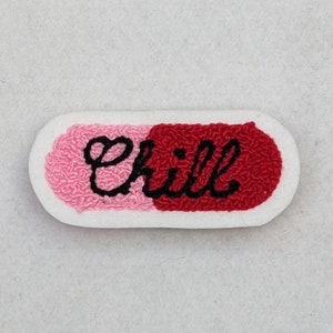 Op de afbeelding: Een witte, pilvormige patch met roze en rode stof. Het woord "chill" is geborduurd in zwarte cursieve letters.