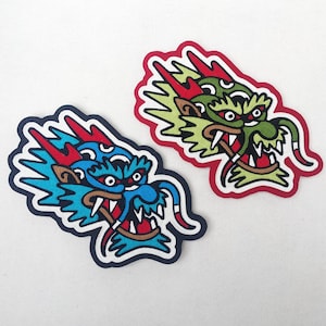 May include: Two embroidered iron-on patches featuring a dragon head design. The dragon on the left is blue and green, while the dragon on the right is green and yellow. Both dragons have red accents.