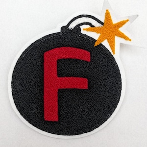 Peut inclure: Écusson brodé représentant une bombe noire avec un "F" rouge au centre. La bombe a une mèche blanche et un motif d'éclat d'étoile jaune. L'écusson est bordé de blanc.