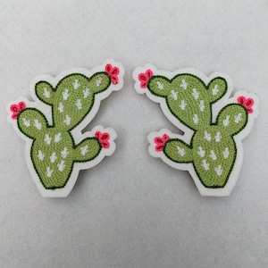 May include: Two green cactus patches with white accents and pink flowers. The patches are embroidered and have a white background.