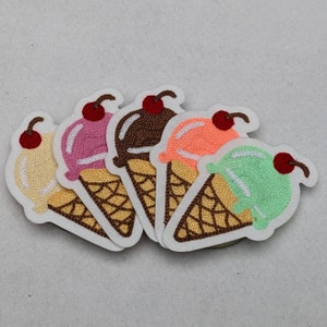 Op de afbeelding: Vijf geborduurde ijsjes in verschillende kleuren: geel, roze, bruin, oranje en groen. Elke patch heeft een wafelkegel en een kers. De patches staan op een witte achtergrond.