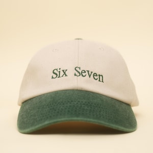Könnte beinhalten: Beige Baseballkappe mit grünem Schirm. Der Text "Six Seven" ist grün auf die Vorderseite gestickt. Der Hut befindet sich vor einem hellgelben Hintergrund.