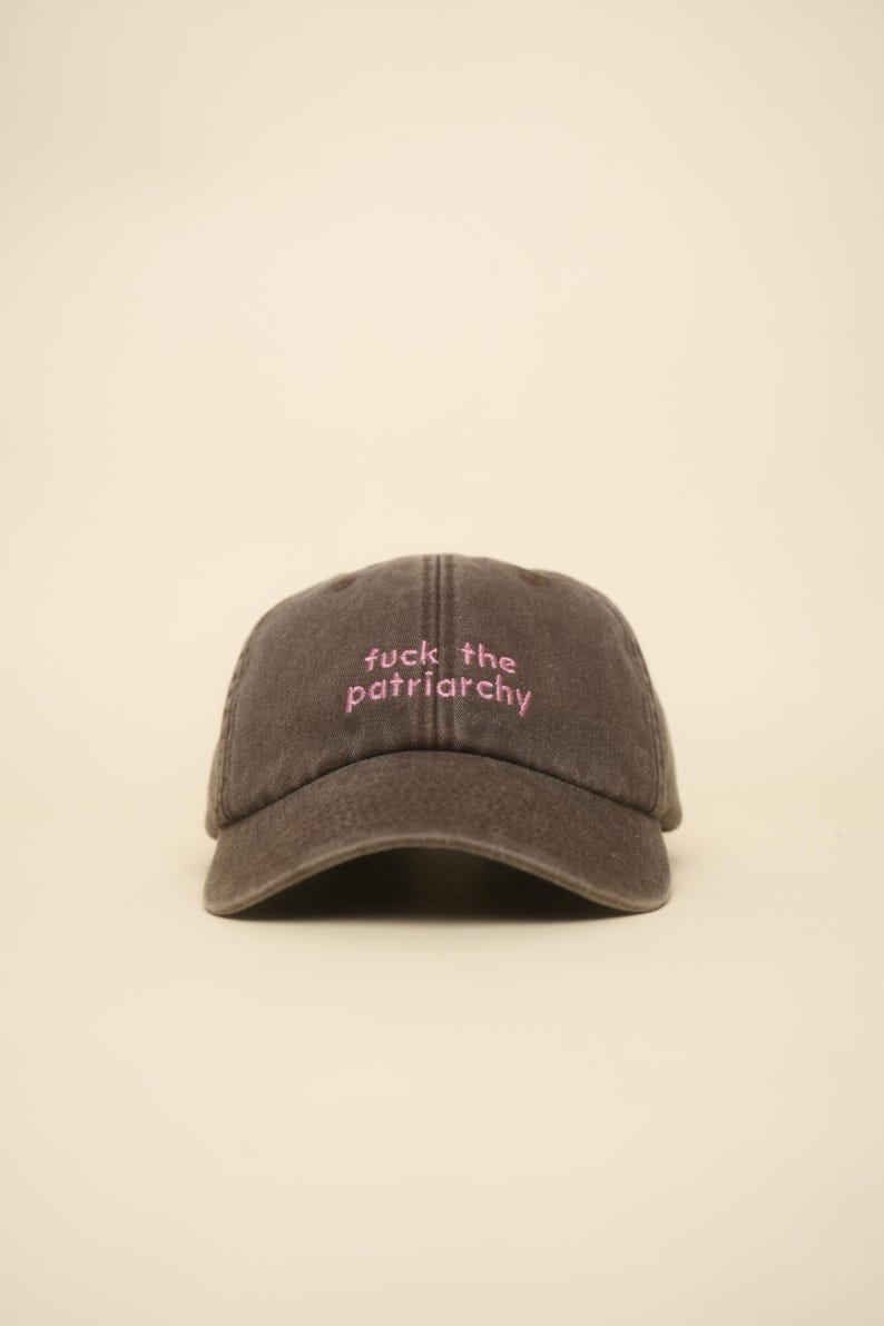 K&ouml;nnte beinhalten: Dunkelbraune Baseballkappe mit dem pinkfarbenen Schriftzug "fuck the patriarchy". Die Kappe hat einen gebogenen Schirm und ein klassisches Sechs-Panel-Design, geeignet f&uuml;r Freizeitkleidung.