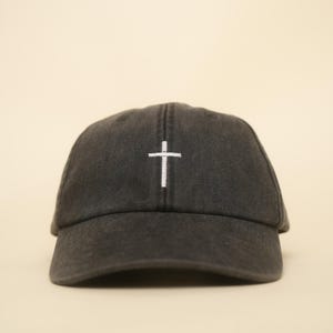 Könnte beinhalten: Dunkelgraue Baseballkappe mit einem weißen, gestickten Kreuz auf der Vorderseite. Die Kappe hat einen gebogenen Schirm und ein klassisches Sechs-Panel-Design. Der Hintergrund ist einfarbig und neutral.