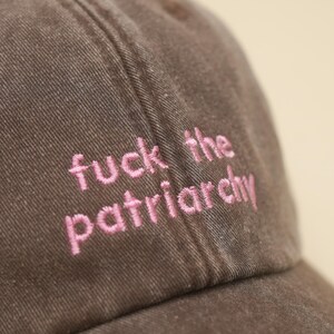 K&ouml;nnte beinhalten: Braune Baseballkappe mit dem in rosa gestickten Schriftzug "fuck the patriarchy". Die Kappe besteht aus einem strukturierten Stoff und hat einen gebogenen Schirm. Der Text ist mittig auf der Vorderseite.