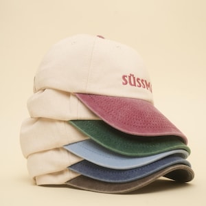 Könnte beinhalten: Ein Stapel Baseballkappen in verschiedenen Farben. Die Kappen haben eine cremefarbene Krone und einen farbigen Schirm. Das Wort "stüssy" ist auf der Vorderseite der Kappe in einer Farbe gestickt, die zum Schirm passt.