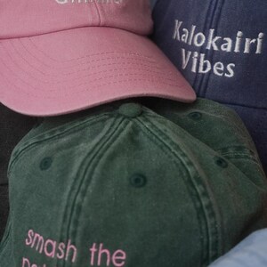 K&ouml;nnte beinhalten: Eine Sammlung von Baseballkappen in verschiedenen Farben, darunter Rosa, Marineblau und Gr&uuml;n. Die rosa Kappe hat dreimal das Wort "Gimme" gestickt. Die marineblaue Kappe tr&auml;gt die Aufschrift "Kalokairi Vibes". Die gr&uuml;ne Kappe tr&auml;gt die Aufschrift "smash the patriarchy".