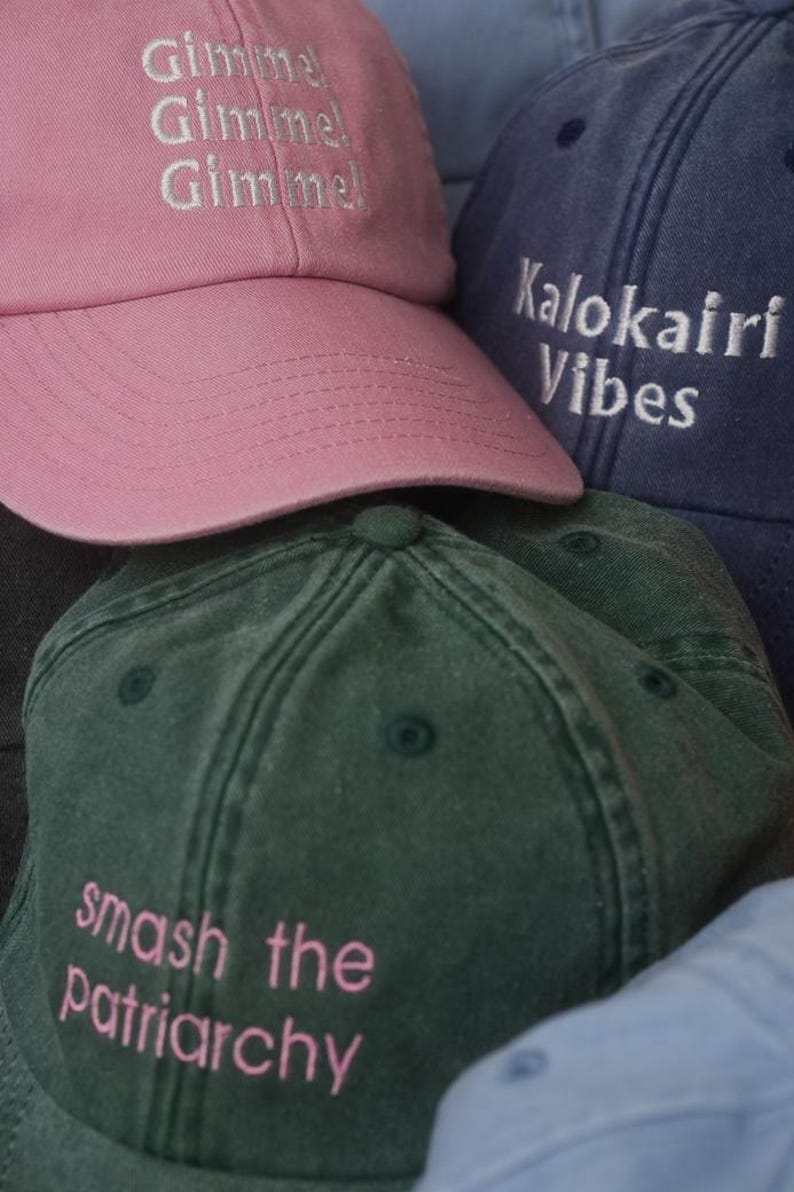 K&ouml;nnte beinhalten: Eine Sammlung von Baseballkappen in verschiedenen Farben, darunter Rosa, Marineblau und Gr&uuml;n. Die rosa Kappe hat dreimal das Wort "Gimme" gestickt. Die marineblaue Kappe tr&auml;gt die Aufschrift "Kalokairi Vibes". Die gr&uuml;ne Kappe tr&auml;gt die Aufschrift "smash the patriarchy".