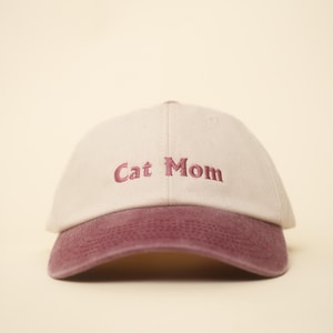 Könnte beinhalten: Beige Baseball Cap mit einem bordeauxfarbenen Schirm. Auf der Vorderseite ist der Schriftzug "Cat Mom" in Bordeauxrot gestickt. Eine lässige Kopfbedeckung.
