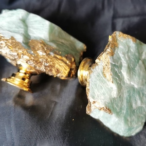 Puede incluir: Un tapón de botella decorativo con dos piezas de piedra verde claro con detalles dorados. El tapón tiene una base de metal dorado. La piedra tiene una textura rugosa y natural.