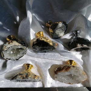 Peut inclure: Plusieurs flacons de parfum décoratifs. Chaque flacon est composé d'un corps en pierre brute unique, orné d'un bouchon doré. Les pierres présentent des couleurs variées, avec des formations cristallines visibles et des accents de feuilles d'or. Les flacons sont disposés sur un tissu blanc.