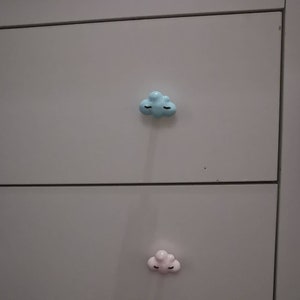 Może przedstawiać: Jasnoszara komoda z dwiema szufladami, każda ozdobiona gałką w kształcie chmury. Górna gałka jest jasnoniebieska, a dolna różowa. Obie gałki mają czarne detale w postaci zamkniętych oczu.