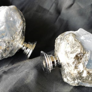 Könnte beinhalten: Zwei dekorative Schrankknöpfe. Jeder Knopf hat einen hellblauen, rau strukturierten Stein, der an einer silberfarbenen Metallbasis befestigt ist. Die Knöpfe werden auf einer dunklen, strukturierten Oberfläche präsentiert.