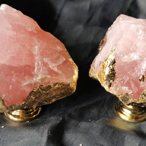 Könnte beinhalten: Zwei Schrankknöpfe aus Rosenquarz mit vergoldeten Kanten und einem goldenen Sockel. Der rosa Stein hat eine raue, natürliche Textur. Die Knöpfe sind vor einem dunklen, strukturierten Hintergrund platziert.