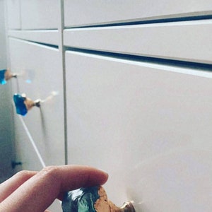 Könnte beinhalten: Nahaufnahme von weißen Schranktüren mit dekorativen blauen und goldenen Schrankknöpfen. Die Knöpfe haben ein einzigartiges, strukturiertes Aussehen und verleihen dem minimalistischen Design einen Hauch von Eleganz. Die Schranktüren sind sauber und modern.