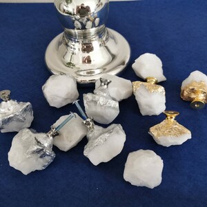 Może przedstawiać: Kolekcja białych kwarcowych gałek do szafek ze srebrnymi i złotymi akcentami. Gałki są przymocowane do kwarcu za pomocą srebrnego osprzętu. Gałki znajdują się na niebieskiej powierzchni.