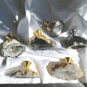 Könnte beinhalten: Sechs dekorative Parfümflakons mit goldfarbenen Verschlüssen und Körpern aus Naturstein. Die Flakons sind auf einer weißen, strukturierten Oberfläche angeordnet. Die Steinkörper weisen eine Mischung aus Grau, Weiß und Braun auf.