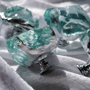 Op de afbeelding: Een verzameling van zes decoratieve kastknoppen. Elke knop heeft een ruwe, turquoise gekleurde glazen bovenkant met een zilverkleurige basis. De knoppen zijn gerangschikt op een wit, getextureerd oppervlak, waarbij hun unieke, organische vormen en metalen accenten worden getoond.