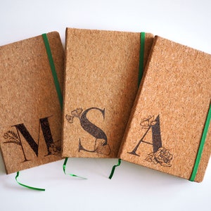 Könnte beinhalten: Drei Kork-Notizbücher mit grünen Bandverschlüssen. Jedes Notizbuch hat einen anderen Buchstaben, M, S und A, mit floralen Designs, die auf die Vorderseite graviert sind.