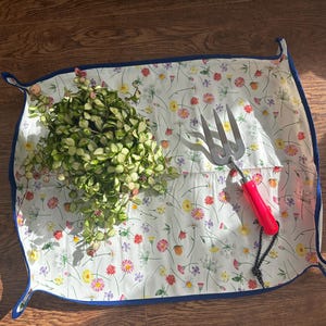 May include: A white floral print gardening mat with a blue border, holding a small plant and a gardening hand fork with a red handle. The mat is filled with colorful flowers and green leaves.