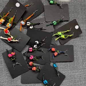 May include: A collection of handmade earrings displayed on black rectangular cards. Each pair features colourful beads wrapped in thin, textured cord or thread, attached to silver-toned metal earring hooks. The designs vary in colour, including orange, red, green, pink, and blue accents.