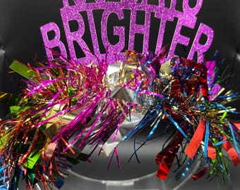 May include: A pink glitter paper crown with the text "100 Days Brighter" on it. The crown is decorated with colorful tinsel streamers.