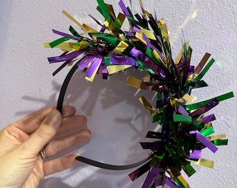 May include: A black headband with a large, colorful bow made of green, gold, and purple metallic streamers.