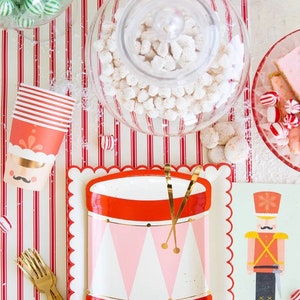 Puede incluir: Platos de papel rojo y blanco con un diseño de tambor y detalles dorados. Los platos están sobre un mantel a rayas rojas y blancas con una ilustración de cascanueces en una tarjeta verde.