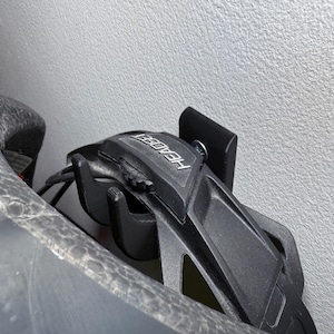Puede incluir: Soporte para casco negro con la palabra "HEADER" impresa en &eacute;l. El soporte est&aacute; montado en una pared blanca.