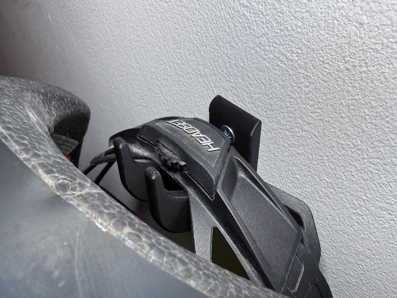 Puede incluir: Soporte para casco negro con la palabra "HEADER" impresa en &eacute;l. El soporte est&aacute; montado en una pared blanca.