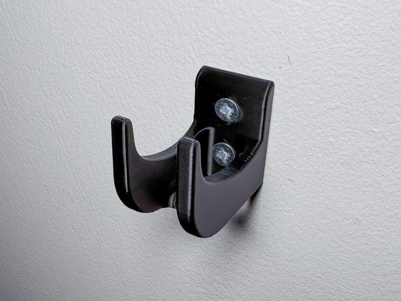 Puede incluir: Un gancho de pared de pl&aacute;stico negro con dos tornillos. El gancho tiene un dise&ntilde;o curvo para sujetar un objeto.