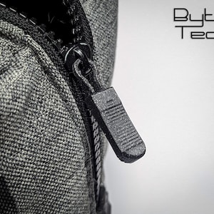 May include: Close-up of a black zipper pull with a textured surface. The zipper pull is attached to a gray fabric zipper.