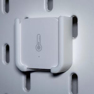 Op de afbeelding: Een wit, vierkant smart home-apparaat met een thermometerpictogram, gemonteerd op een wit pegboard. Het apparaat wordt op zijn plaats gehouden door een witte plastic beugel. Het pegboard heeft gelijkmatig verdeelde ovale gaten.
