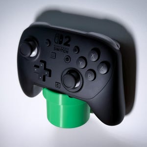 May include: A black Nintendo Switch Pro Controller on a green cylindrical holder. The controller features the Nintendo Switch logo and button labels. The holder is designed to look like a green pipe, often seen in video games.