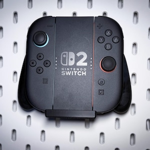 May include: A black Nintendo Switch controller with the text "Nintendo Switch" and the number "2" printed on the front. The controller has buttons in red, blue, and black. The controller is on a white surface with evenly spaced holes.