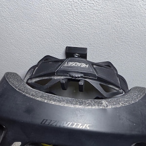Puede incluir: Un casco de bicicleta Specialized negro con un soporte de casco negro fijado a una pared blanca. El soporte de casco tiene la palabra "HEADSET" impresa.